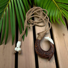 Wooden fish hook pendant with rope on a wooden surface with green leaves in the background