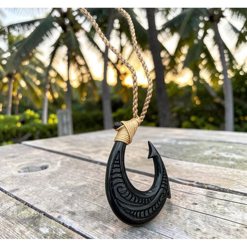 A carved black fishhook pendant with tribal-style engravings, wrapped near the top with tan cord, hanging on a braided tan rope against a blurred tropical outdoor background with palm trees and warm sunset light.