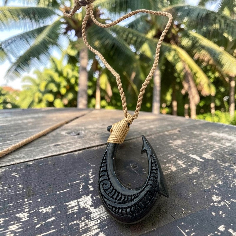 A carved black bone or wood Polynesian-style fishhook pendant necklace with a natural tan cord, resting on a weathered wooden table with a tropical palm forest background.