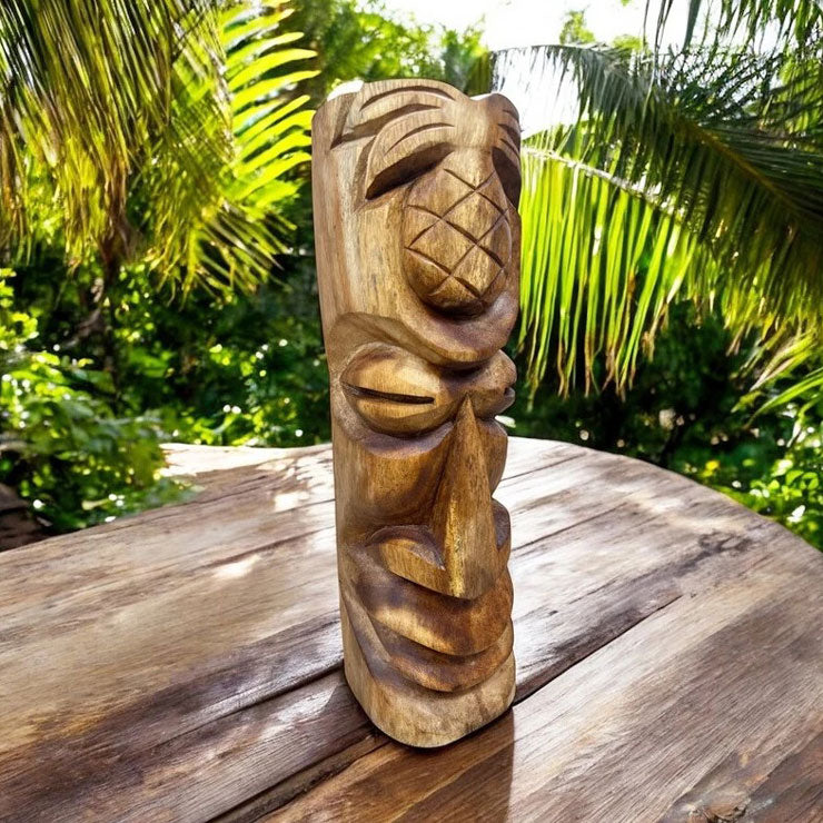 A hand-carved wooden tiki totem sculpture standing on a rustic wooden table, with a lush green tropical garden background.