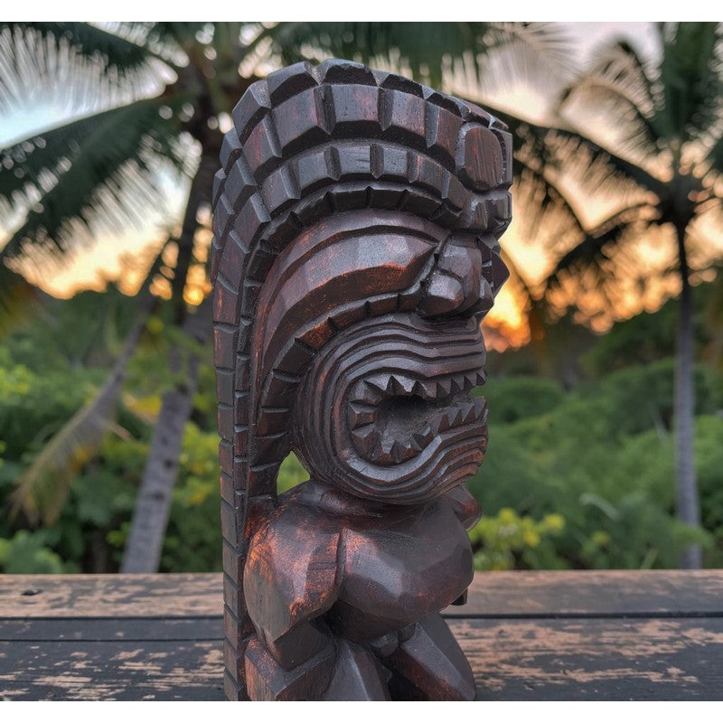 A dark carved wooden tiki statue with a detailed headdress and open mouth, set outdoors on a wooden surface with palm trees and a sunset sky in the background.