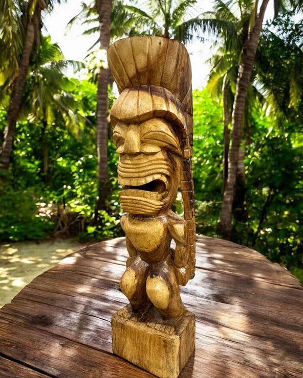 A hand-carved wooden Kanaloa tiki statue with a detailed face and crown, displayed on a wooden table with a lush tropical garden background.