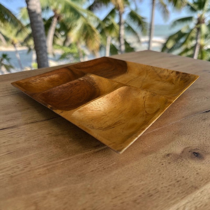 A rectangular wooden divided tray with three compartments, resting on a wooden table with a blurred tropical beach and palm tree background.