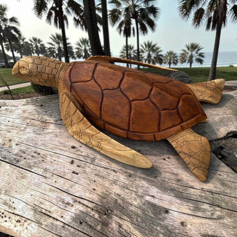 A carved wooden sea turtle bag with a shoulder strap, resting on a weathered wooden surface with a beach and palm trees in the background.
