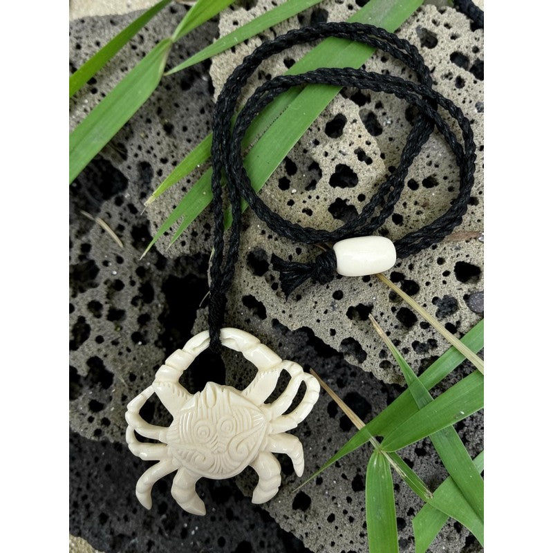 White crab-shaped pendant on a black cord with green leaves and stone background