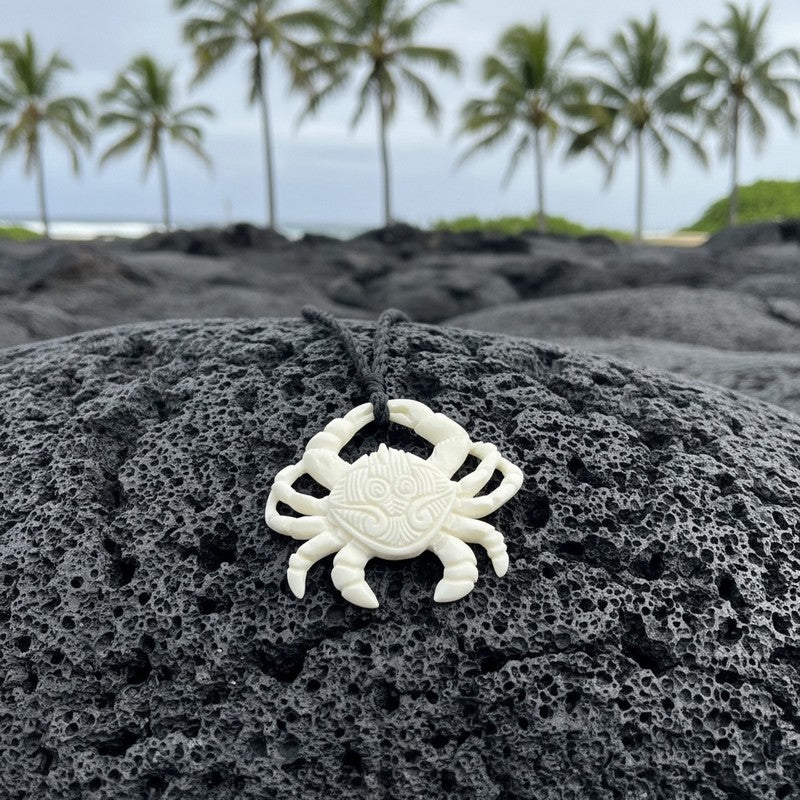 White crab pendant on a black cord against a backdrop of black volcanic rock and palm trees.