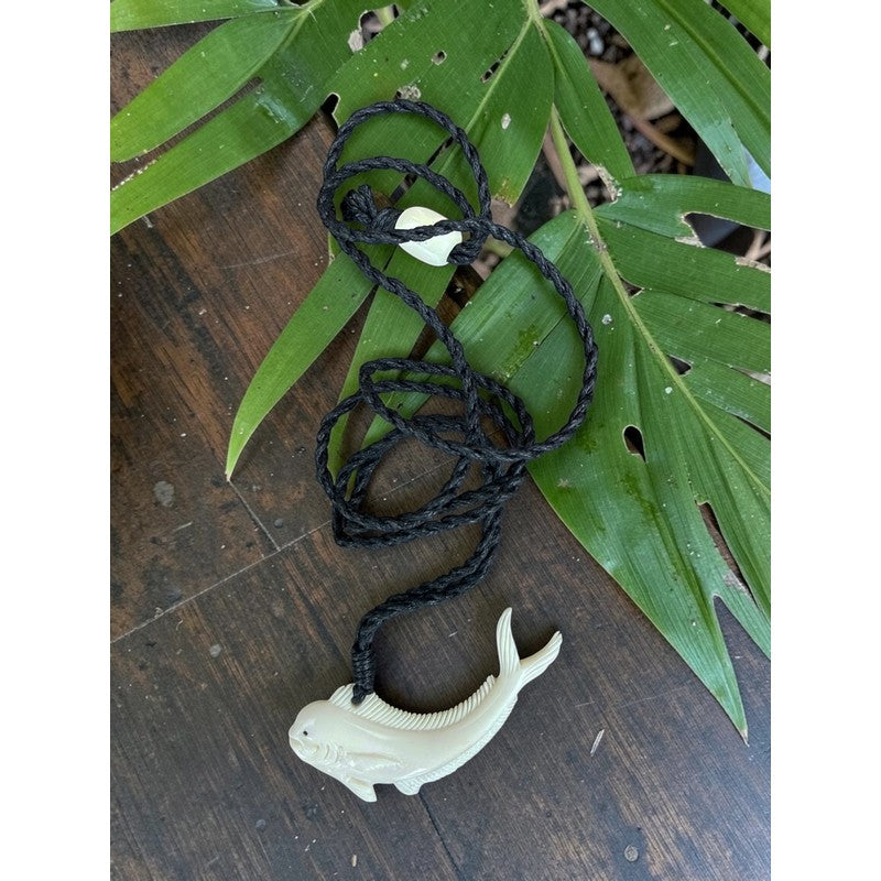 White fish-shaped pendant on a black cord with green leaves on a wooden surface