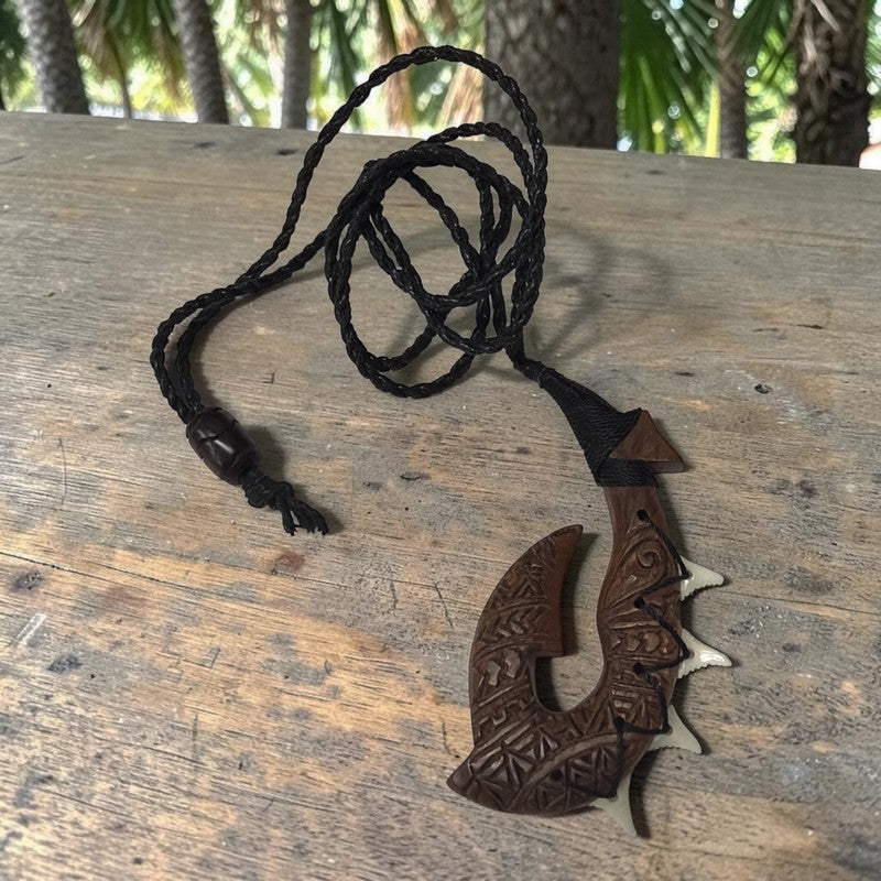 A carved wooden pendant necklace with a black braided cord, displayed on a rustic wooden table with a blurred outdoor background of palm trees.