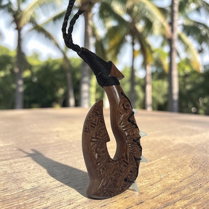 A carved wooden Māori-style hook pendant with a black braided cord, photographed standing upright on a wooden surface with palm trees blurred in the background.