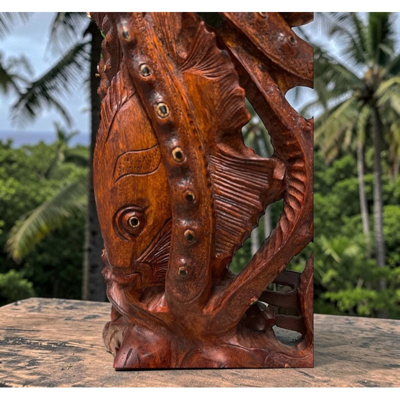 A hand-carved wooden fish sculpture with a warm reddish-brown finish, displayed on a stone surface with a blurred tropical garden background.