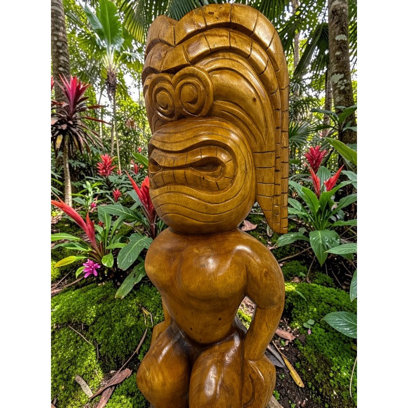 Wooden tiki statue in a tropical forest setting with colorful flowers and greenery.