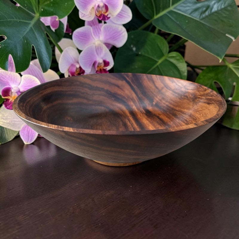 A round wooden bowl with a rich dark brown and black grain pattern, set on a dark wooden table with purple orchids and green leaves in the background.