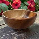 A round wooden bowl with a smooth, polished finish sits on a weathered dark wooden table, with a blurred background of green leaves and red hibiscus flowers.