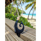 Black fish hook pendant on a wooden surface with a tropical beach background