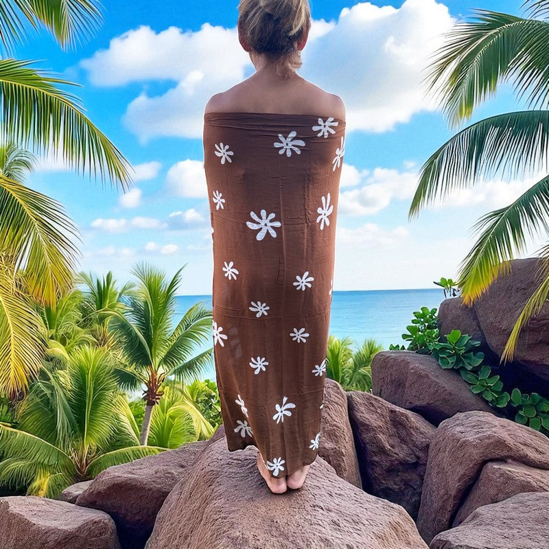 Person wearing a brown floral dress standing on rocks with palm trees and ocean in the background