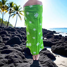 Person wearing a green floral sarong on a rocky beach with palm trees and ocean in the background.
