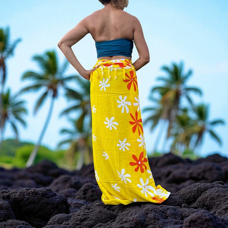 Person wearing a yellow floral skirt on a rocky beach with palm trees in the background