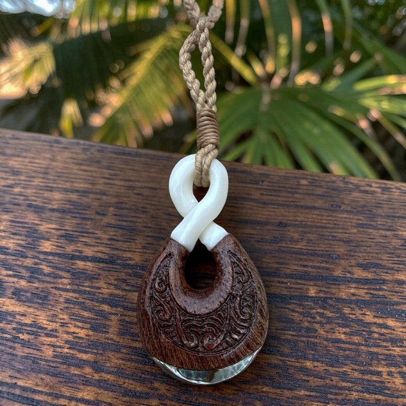 A carved wooden pendant with a white twisted cord loop and a clear glass disc at the bottom, resting on a dark wooden surface with blurred green foliage in the background.
