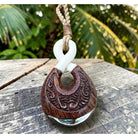 A carved wooden pendant with a white loop and a natural fiber cord, resting on a weathered wooden surface with a blurred tropical green background.