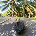 A carved black bone or wood Polynesian-style fishhook pendant necklace with a natural tan cord, resting on a weathered wooden table with a tropical palm forest background.