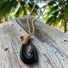 A black carved fishhook pendant necklace with a natural tan braided cord, resting on a weathered wooden surface with a blurred tropical palm forest background.