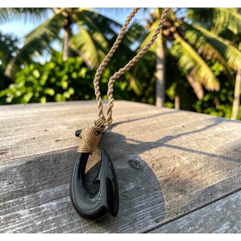 A black carved fishhook pendant necklace with a natural tan rope cord, resting on a weathered wooden surface with a tropical palm tree background.