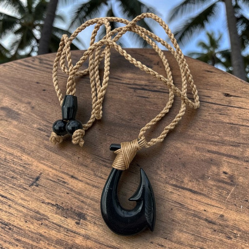 A black carved fishhook pendant necklace with a tan braided cord, resting on a rustic wooden surface with blurred palm trees in the background.