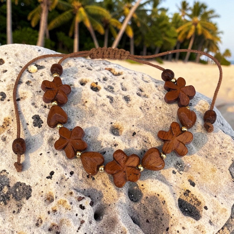 A brown cord bracelet with carved wooden flower and heart beads, displayed on a textured rock with a beach and palm trees in the background.