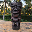 A dark brown carved wooden tiki statue with a fierce, toothy grin, standing on a weathered wooden surface with a tropical garden background featuring palm trees.