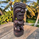 A dark carved wooden tiki statue with a fierce face and headdress, standing on a weathered wooden table with a tropical garden and palm trees in the background.