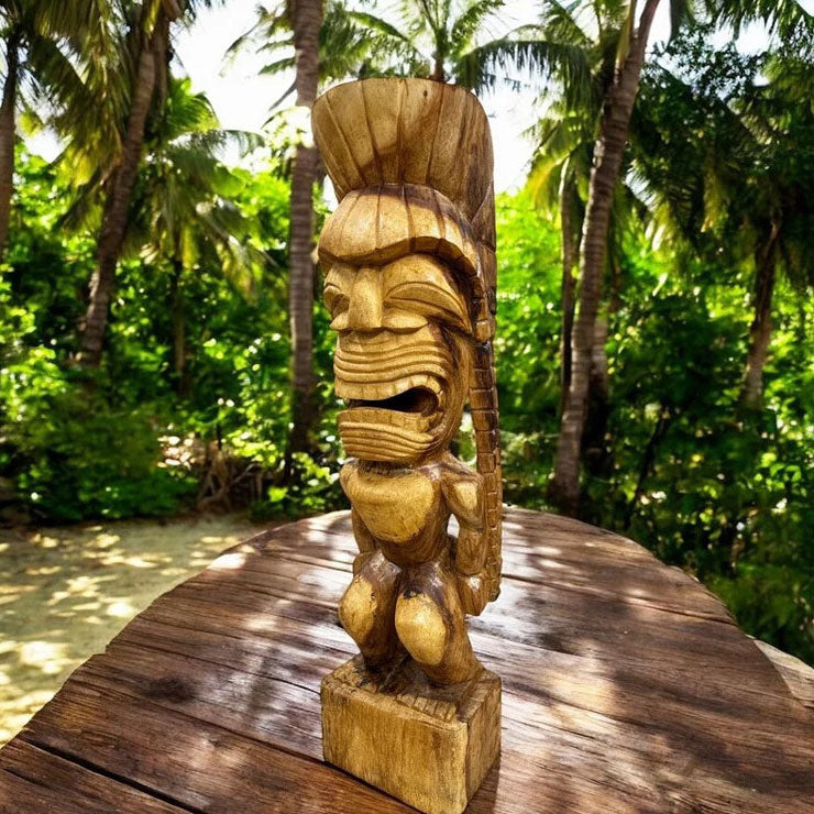 A hand-carved wooden Kanaloa tiki statue with a detailed face and crown, displayed on a wooden table with a lush tropical garden background.