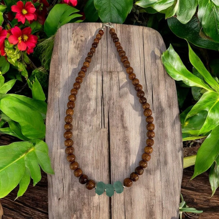 Brown beaded necklace with a green leaf pendant on a wooden surface surrounded by greenery