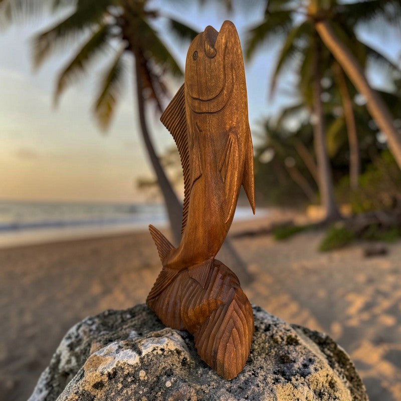 A carved wooden fish sculpture standing upright on a rock, photographed outdoors on a beach with palm trees and the ocean blurred in the background.