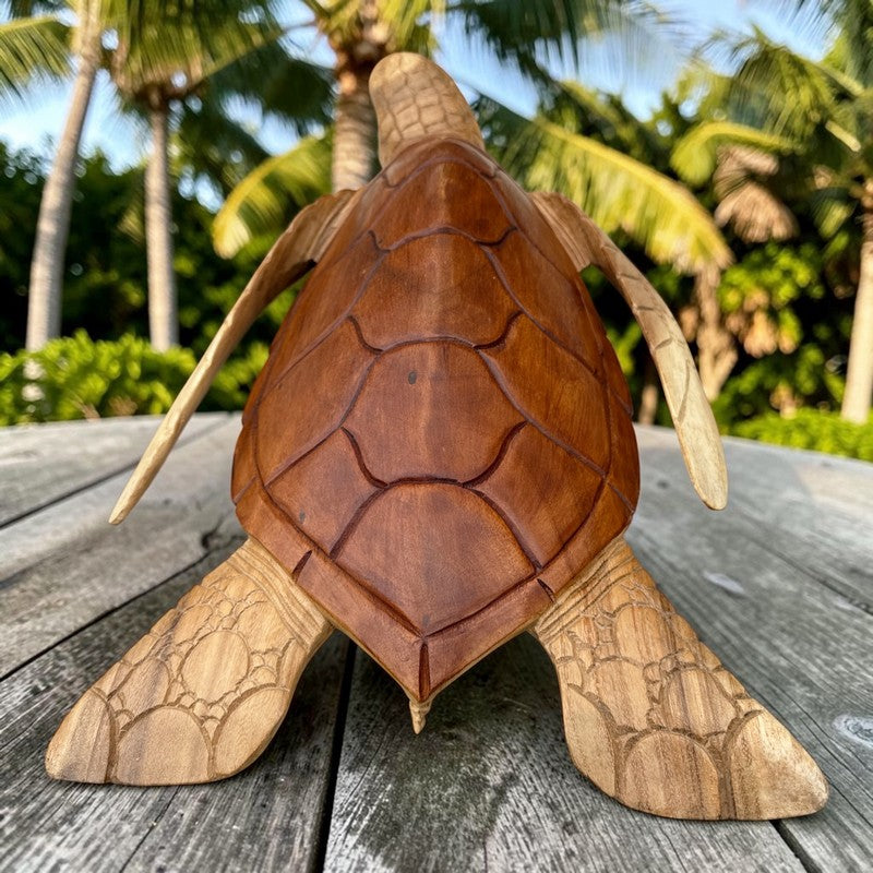 A handcrafted wooden sea turtle sculpture displayed on a weathered gray wooden table, with a blurred background of palm trees and green foliage.