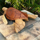 A hand-carved wooden sea turtle sculpture resting on a weathered wooden deck with green foliage in the background.