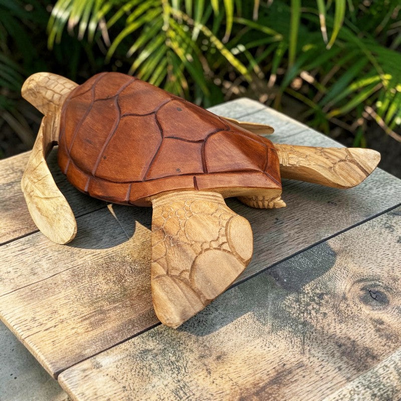 A hand-carved wooden sea turtle sculpture resting on a weathered wooden deck with green foliage in the background.