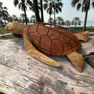 A carved wooden sea turtle bag with a shoulder strap, resting on a weathered wooden surface with a beach and palm trees in the background.