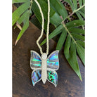Butterfly-shaped pendant with abalone shell wings on a string, placed on a wooden surface with leaves in the background.