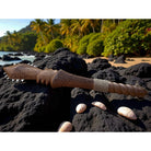 Decorative wooden staff with carvings on a black volcanic rock surface with a tropical beach background.