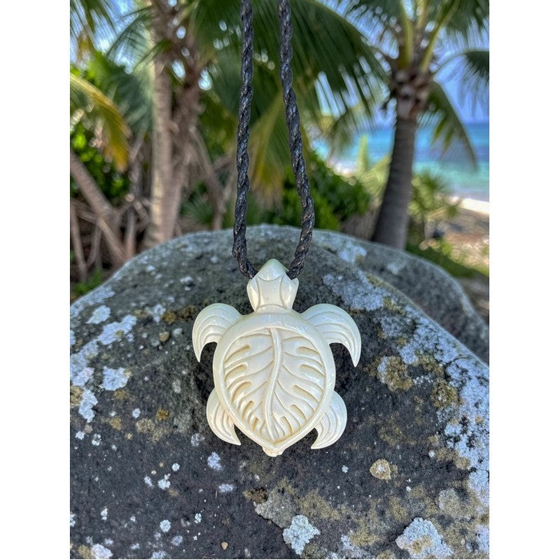 Turtle pendant on a rope against a natural background with palm trees and rocks.