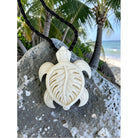 Carved turtle pendant on a rope against a beach backdrop with palm trees and ocean.
