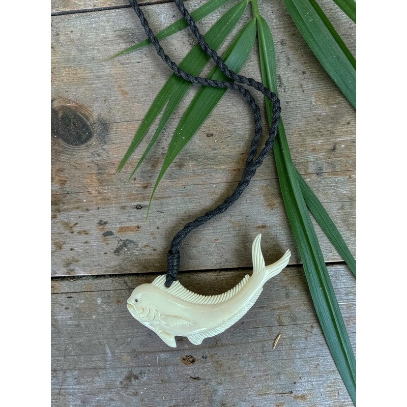White fish-shaped pendant on a black cord with green leaves on a wooden surface