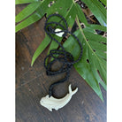 White fish-shaped pendant on a black cord with green leaves on a wooden surface