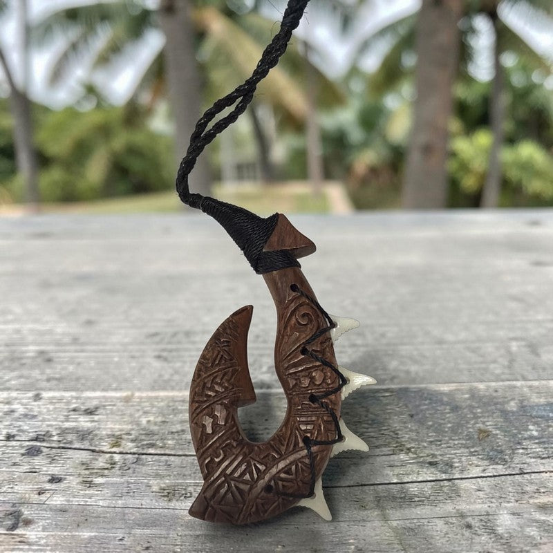A carved wooden and bone fishhook pendant necklace with a black braided cord, resting on a weathered gray wooden surface with a blurred tropical outdoor background.