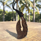 A carved wooden Māori-style hook pendant with a black braided cord, photographed standing upright on a wooden surface with palm trees blurred in the background.