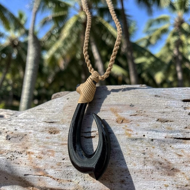 A black carved fishhook pendant necklace with a tan braided cord, resting on a weathered wooden log with a tropical background of palm trees and blue sky.