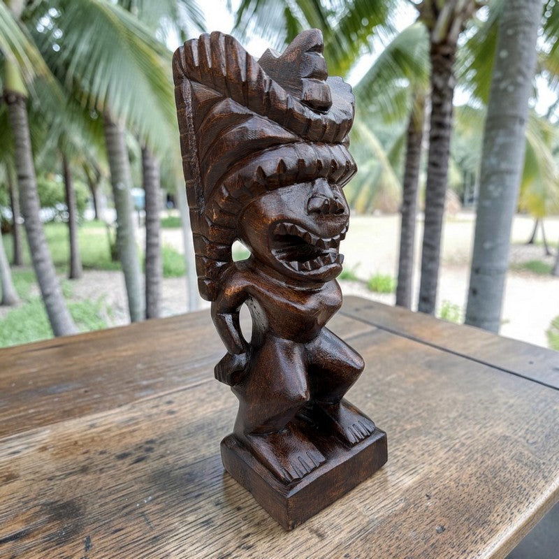 A dark brown carved wooden tiki statue of a stylized figure with a large headdress, displayed on a rustic wooden table with a blurred tropical background of palm trees.