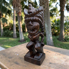 A dark brown carved wooden tiki statue with a wide-brimmed feathered headdress, standing on a wooden table with a blurred tropical garden background.