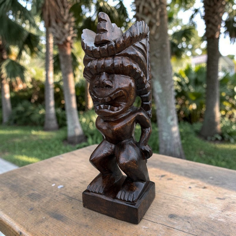 A dark brown carved wooden tiki statue with a wide-brimmed feathered headdress, standing on a wooden table with a blurred tropical garden background.