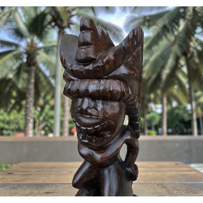 A dark brown carved wooden tiki statue with a detailed headdress, displayed outdoors on a wooden surface with a blurred background of palm trees.
